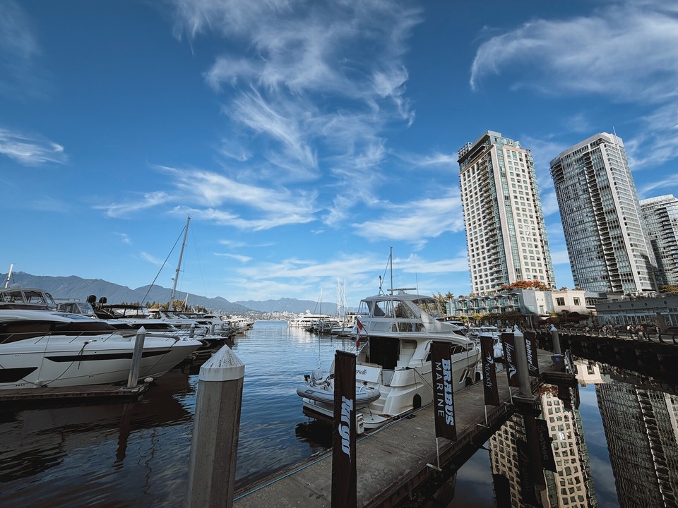 Boats moored in a marina beside waterfront towers, with mountains beneath a blue sky